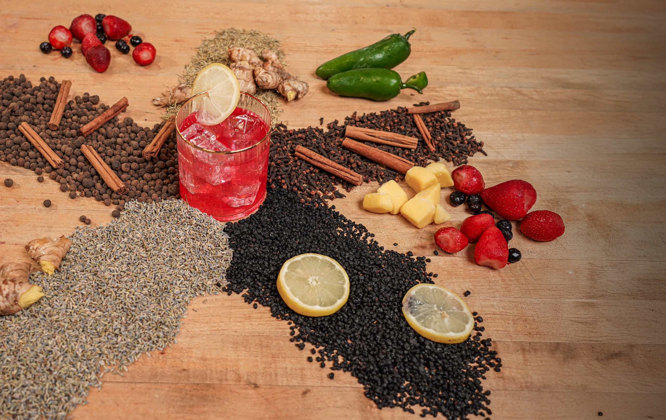 Glass of red kombucha on a wooden surface surrounded by fresh ingredients including ginger, lemon slices, cinnamon sticks, elderberries, lavender, and strawberries — representing natural kombucha flavors from Firefly Brew.