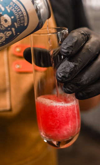 Pouring Blueberry Lemonade kombucha into a glass — artisan brewer in brown apron pouring Firefly Booch Brew’s vibrant pink kombucha made with organic ingredients.